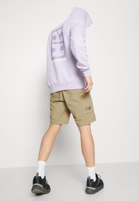Licht lavendelkleurige hoodie met een groot logo op de rug, gecombineerd met beige shorts met een zijzak. Zwarte schoenen en witte sokken.