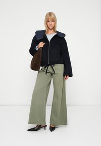 Chaqueta de punto azul marino, pantalones anchos verdes oliva y zapatos negros de punta. El modelo sostiene un bolso de ante marrón. Fondo liso.