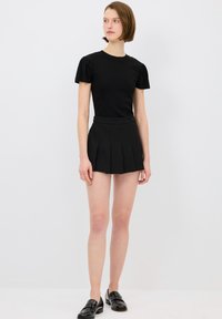 Černé úzké tričko s krátkým rukávem a nabíranými rukávy je zkombinováno s černou skládací sukní. Lesklé černé mokasíny outfit dokončují.