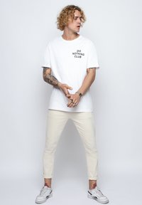 White cotton t-shirt with "DO NOTHING CLUB" print, paired with cream-colored cuffed pants and white sneakers. Skin visible on arms with tattoos.