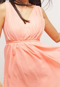 Svetle broskvové šaty bez rukávů s výstřihem do V, skládáním v látce, nařaseným pasem a detailním pleteným pásem pro přidanou texturu a design.