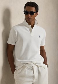 Mężczyzna w białej koszulce polo z podniesionym kołnierzykiem, białych spodniach, ciemnych okularach przeciwsłonecznych, stojący z rękami w kieszeniach na tle neutralnej ściany.