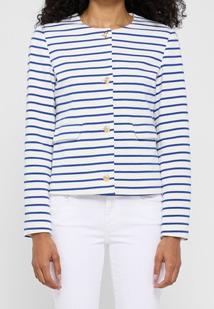 Mujer con chaqueta blanca con rayas horizontales azules y botones dorados, combinada con pantalones blancos, mostrada desde el cuello hasta las caderas.