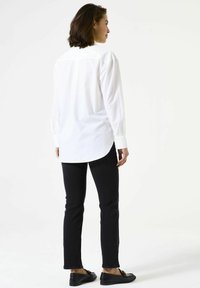 Chemise blanche à boutons avec manches longues, coupe droite et ourlet incurvé, portée avec un jean skinny noir et des chaussures plates noires.