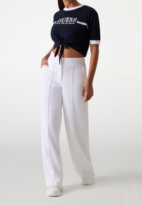 Navy cropped t-shirt with a knotted front and white logo, paired with high-waisted white wide-leg trousers and white sneakers.