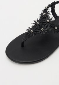 Svart tøflesandal med blomsterdekorasjoner og sølvperler. Glatt materiale med en flat, rund såle.
