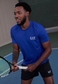 Camisa atlética azul de manga corta, con logo en el pecho, y pantalones cortos negros. Sosteniendo una raqueta de tenis con cabeza verde y negra, en una cancha.