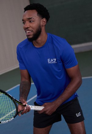 Blue athletic shirt with short sleeves, logo on chest, and black shorts. Holding a tennis racket with a green and black head, on a court.