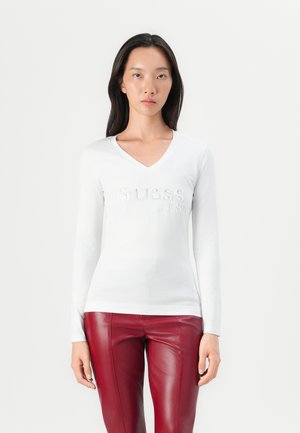T-shirt blanc à manches longues en V, avec "GUESS est. 1981" en strass, associé à un pantalon en cuir synthétique rouge, porté par un mannequin.