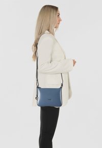 Borsa a tracolla in pelle blu con texture liscia, tracolla regolabile, ferramenta argentata e chiusura con zip, indossata sulla spalla.