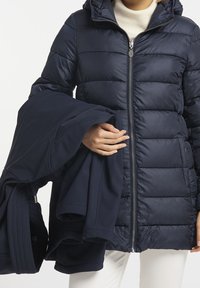 Chaqueta acolchada de color marino con cremallera frontal y capucha, que presenta un diseño acolchado. En la otra mano se sostiene una prenda a juego de color marino.