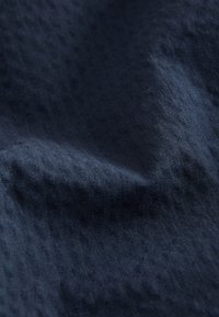 Tamsiai mėlynos tekstūruotos audinio artimas planas su subtiliomis horizontaliomis raštais ir švelniais klostėmis, kurios sukuria šešėlius ir šviesos atspindžius.