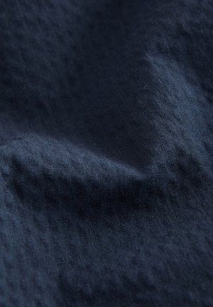 Lähikuva tumma sinisestä teksturoidusta kankaasta, jossa on hienovaraisia vaakasuoria kuvioita ja pehmeitä laskoksia, jotka luovat varjoja ja korostuksia.