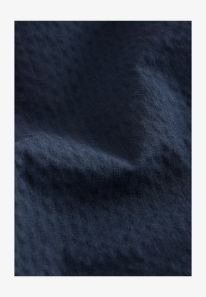 Primo piano di tessuto blu scuro con trama testurizzata, sottili motivi orizzontali e pieghe leggere che creano ombre e luci.