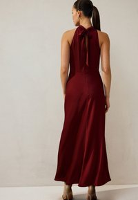 Karmesinrød halter-jumpsuit med en knyttet detalje ved halsen, der har en tætsiddende overdel, der udvider sig til brede ben. Glat stof med en subtil tekstur.