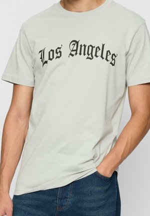 Maglietta grigio chiaro con testo nero in stile Old English "Los Angeles" indossata da una persona con la mano nella tasca dei jeans blu.