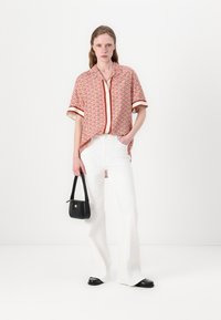 Korte mouw shirt met een geometrisch rood patroon, gedragen over een crème top, gecombineerd met witte broek met wijde pijpen en een zwarte handtas.