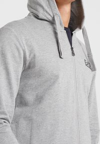 Sudadera gris con cremallera y acabado texturizado, con capucha ajustable y bolsillos laterales; logo visible en el pecho.