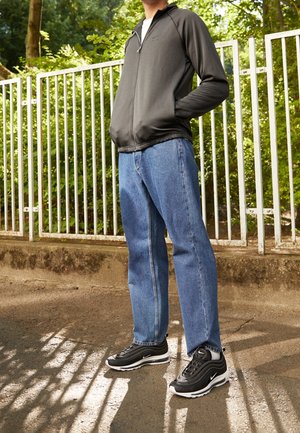 Person steht auf dem Bürgersteig, trägt eine schwarze Zip-Up-Jacke, blaue Jeans und schwarz-weiße Nike-Sneaker, im Hintergrund ein Metallzaun und Grün.