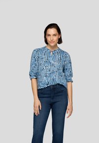 Blau gemusterte Bluse mit Rundhalsausschnitt und Schlitzdetail, verziert mit einer Mischung aus kreisförmigen Mustern. Kombiniert mit dunkelblauen Jeans.