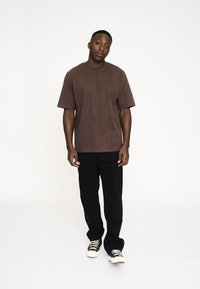 Brun kortærmet T-shirt med en afslappet pasform, der har et lille sort logo på brystet. Kombineret med sorte bukser og beige sneakers.