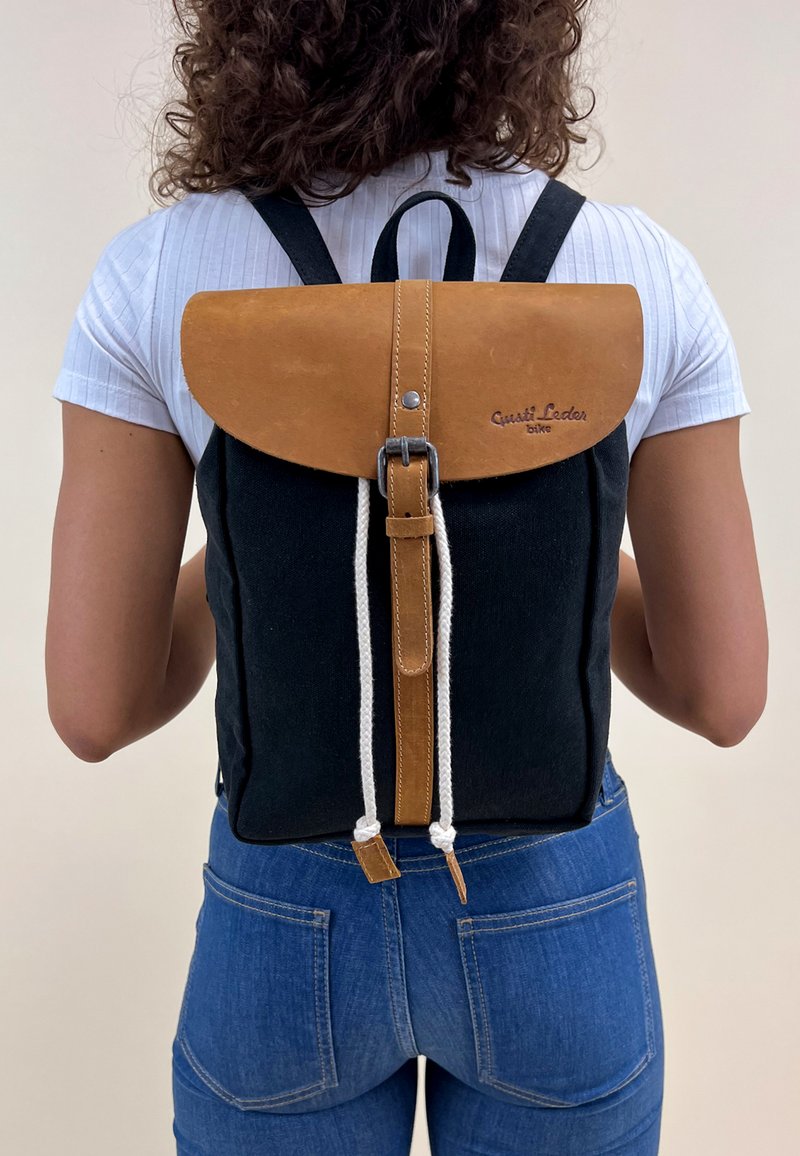 Mochila negra de lona con solapa de cuero marrón, cordón de rope blanco y herrajes metálicos. Presenta un diseño limpio y minimalista.