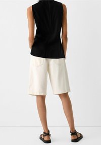 Svart ärmlös topp i kombination med knälånga krämfärgade shorts, med bakfickor, och svarta remsandaler på bara fötter.