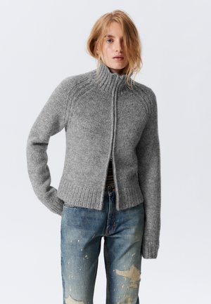 Grå zip-up tröja med hög krage, strukturerad stickning, ribbad nederkant och långa ärmar, samsas med slitna blå jeans.