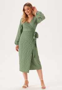 Groene wikkeljurk van textuurstof, met lange mouwen en een striktaille. Heeft een midi-lengte zoom en een subtiel kreukpatroon.