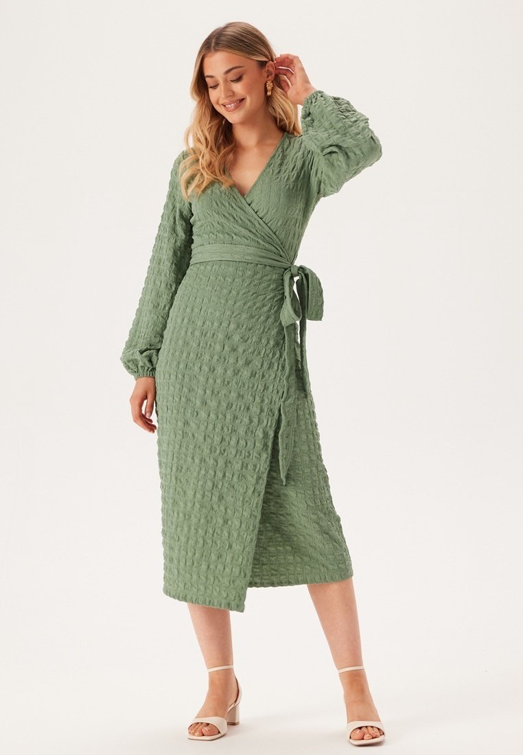 Groene wikkeljurk van textuurstof, met lange mouwen en een striktaille. Heeft een midi-lengte zoom en een subtiel kreukpatroon.