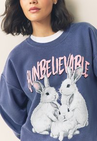 Navyblauwe sweatshirt met een afbeelding van drie witte konijnen en de tekst "BUN BELIEVABLE" in roze, gemaakt van een zachte, gestructureerde stof.
