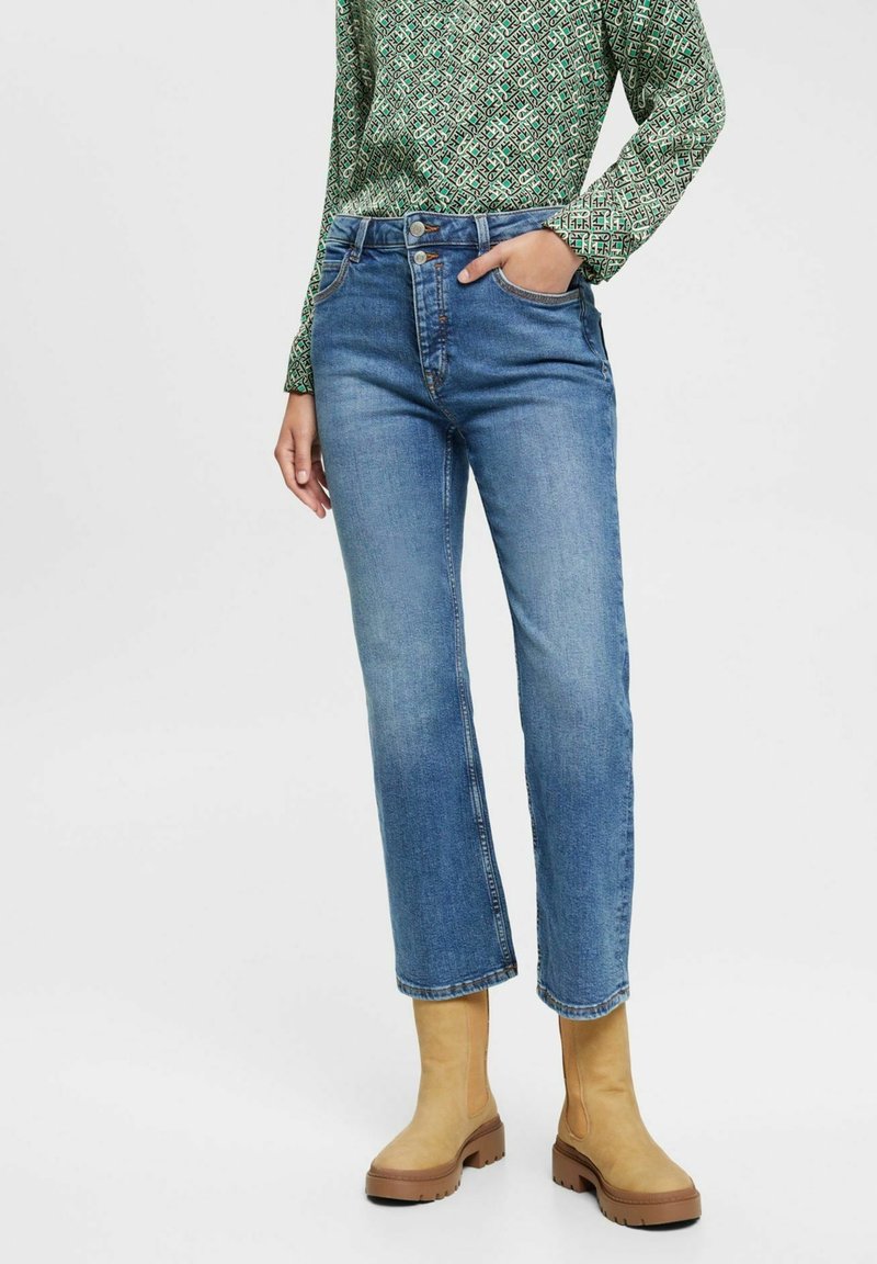 Femme portant un jean bleu à jambes droites, un haut à manches longues vert à motifs, et des bottines épaisses beiges, debout avec une main dans la poche.
