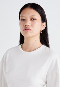 Witte lange mouw shirt met een ronde halslijn, gemaakt van zachte stof, met verlaagde schouders. Gouden hoepel oorbellen zichtbaar. Gladde textuur.