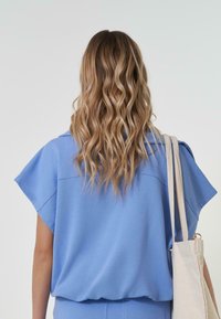 Gaiši zils īsām piedurknēm tops ar brīvu piegriezumu un gludu tekstūru, ar nemanāmu aizmugures šuvi un modeli, kas tur neitrāla toņa somu.