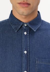 Donkerblauw denim overhemd met knoopsluiting en een klassieke kraag, voorzien van witte knopen en een borstzak. Gladde textuur en stevig materiaal.