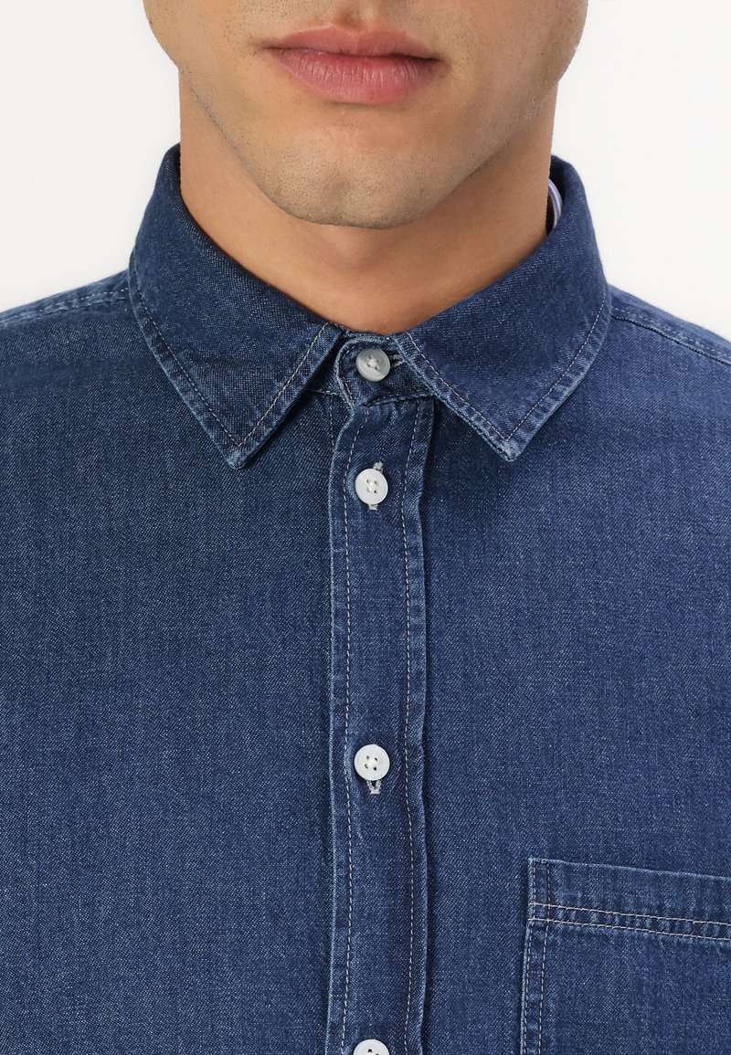 Dunkelblaue Jeanshemd mit klassischem Kragen, weißen Knöpfen und einer Brusttasche. Glatte Textur und strapazierfähiger Stoff.