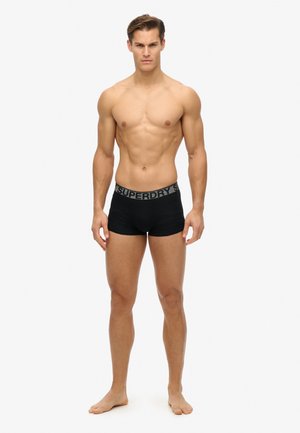 Czarne bokserki z szarym pasem z logo "SUPERDRY". Materiał wydaje się gładki, dopasowany krój. Model stoi boso, z rękami swobodnie opartymi wzdłuż ciała.