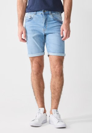 Homme portant un short en jean bleu clair retroussé, une chemise bleu marine et des baskets blanches, debout devant un fond uni.