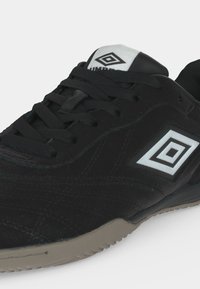 Μαύρο αθλητικό παπούτσι Umbro με λευκό λογότυπο στο πλάι και στη γλώσσα, μαύρα κορδόνια και ανοιχτό γκρι λαστιχένια σόλα.