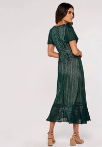 Dunkelgrünes Spitzen-Midikleid mit kurzen Ärmeln, aufwendigen Blattmustern und einem gewellten Saum. Kombiniert mit hellbeigen Blockabsatzsandalen.