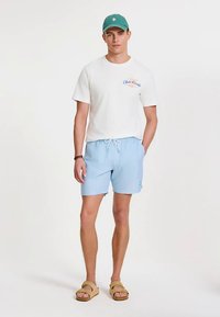 Valkoinen puuvillainen t-paita, jossa teksti "Club Tropique", vaaleansiniset raidalliset uimashortsit kuminauhavyötäröllä ja ruskeat sandaalit. Päällä vihreä lippalakki.