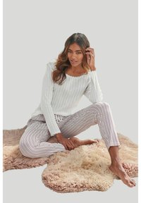Top bianco a maniche lunghe con collo a scallop, abbinato a pantaloni a righe rosa chiaro e bianchi. Modella seduta su tappeti soffici.