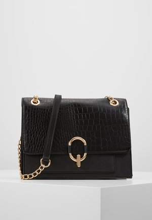 Musta krokotiilikuvioitu nahkalaukku, jossa on kullanvärinen ketjuhihna ja pyöreä kullanvärinen solki, esillä valkoisella jalustalla yksinkertaista harmaata taustaa vasten.