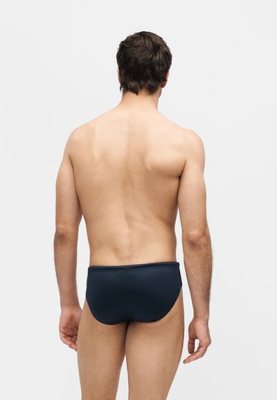 Άνδρας με την πλάτη γυρισμένη, φοράει σκουρόχρωμο μπλε μαγιό μπροστά σε απλό φωτεινό φόντο.