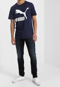Camiseta de algodón azul marino con un gran logo blanco de Puma, combinada con jeans oscuros y zapatillas blancas.