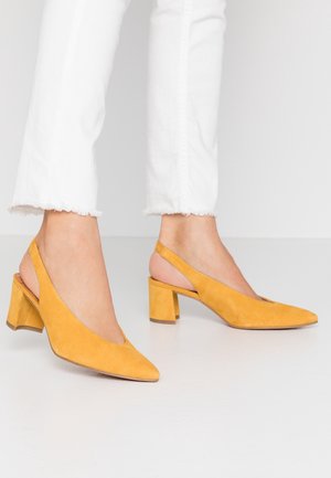 Osoba ubrana w białe krótkie spodnie i żółte zamszowe buty na obcasie typu slingback stoi na białej podłodze.
