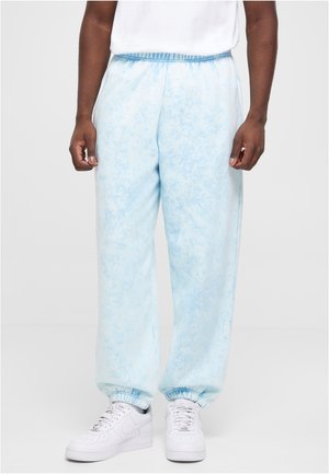 Mand iført lyseblå tie-dye joggingbukser med elastiske manchetter og hvide sneakers, stående mod en ensfarvet baggrund.