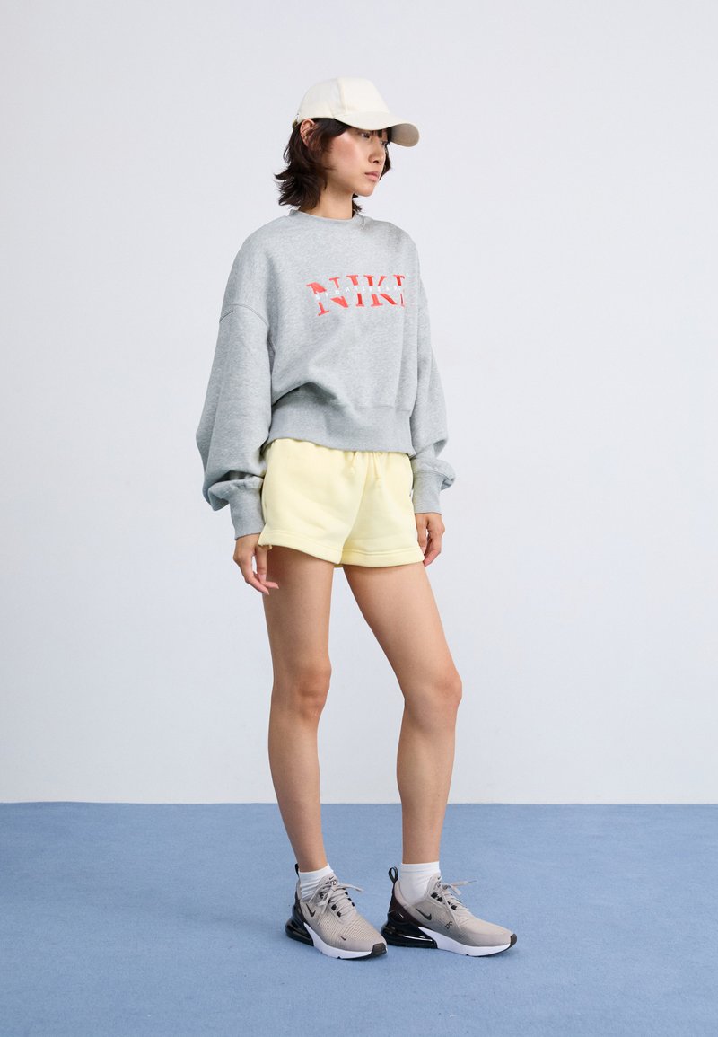 Sudadera gris con logo rojo, pantalones cortos amarillos y zapatillas grises. El modelo lleva una gorra clara; el entorno tiene una alfombra azul y un fondo sencillo.