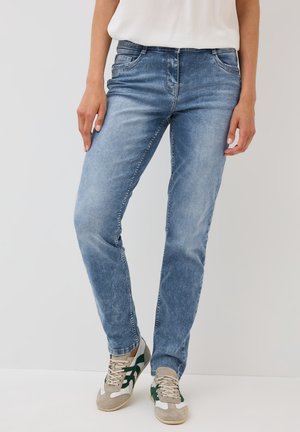 Mujer vestida con jeans claros desgastados y zapatillas beige con acentos en verde oscuro, de pie contra un fondo claro y liso.