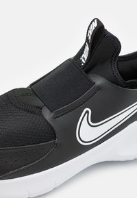 Black athletic shoe with textile upper and elastic strap. Features a prominent white Nike logo and a cushioned white sole.